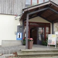 Touristinformation Gohrisch Eingang einer Touristinformation mit Überdachung, Blumenkübeln und Infotafel; das Gebäude hat eine helle Fassade und Holzverkleidung.Entrance to a tourist information center with canopy, flower pots and information board; the building has a light-colored façade and wooden cladding.Vchod do turistického informačního centra s přístřeškem, květináči a informační tabulí; budova má světlou fasádu a dřevěné obložení.Wejście do centrum informacji turystycznej z zadaszeniem, donicami z kwiatami i tablicą informacyjną; budynek ma jasną fasadę i drewnianą okładzinę.Ingang van een toeristisch informatiecentrum met luifel, bloempotten en informatiebord; het gebouw heeft een lichtgekleurde gevel en houten bekleding.Ingresso di un centro di informazione turistica con tettoia, vasi di fiori e pannello informativo; l'edificio ha una facciata chiara e un rivestimento in legno.