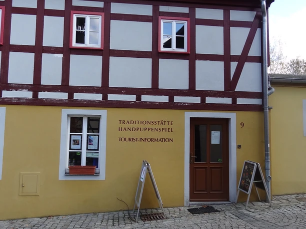 Vakwerkhuis met gele gevel, rode balken en witte ramen; opschrift "Traditionsstätte Handpuppenspiel Tourist-Information" naast houten deur.