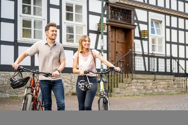 Werther-Innenstadt-Teutoburger-Wald-Tourismus-D-Ketz-038.jpg Zwei Personen mit Fahrrädern stehen lächelnd vor einem Fachwerkhaus in städtischer Umgebung.