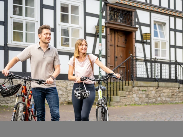 Werther-Innenstadt-Teutoburger-Wald-Tourismus-D-Ketz-038.jpg Zwei Personen mit Fahrrädern stehen lächelnd vor einem Fachwerkhaus in städtischer Umgebung.