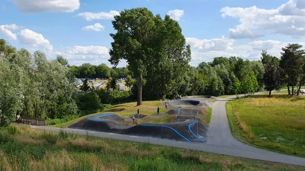 Eine grüne Landschaft mit einem hügeligen Pumptrack, umgeben von Bäumen und Menschen, die Rad fahren.
