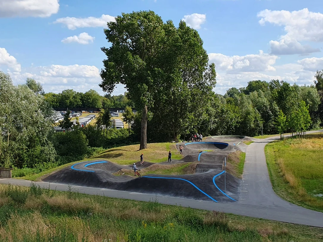 Pumptrack Rheda-Wiedenbrück Eine grüne Landschaft mit einem hügeligen Pumptrack, umgeben von Bäumen und Menschen, die Rad fahren.