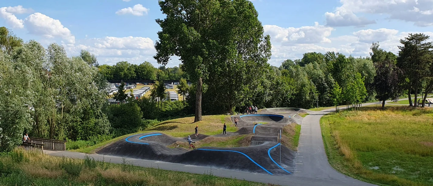 Pumptrack Rheda-Wiedenbrück Eine grüne Landschaft mit einem hügeligen Pumptrack, umgeben von Bäumen und Menschen, die Rad fahren.