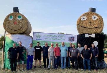 IMG_8992.jpg Eine Gruppe von fröhlichen Menschen posiert vor übergroßen Strohpuppen auf einem Schützenfest.