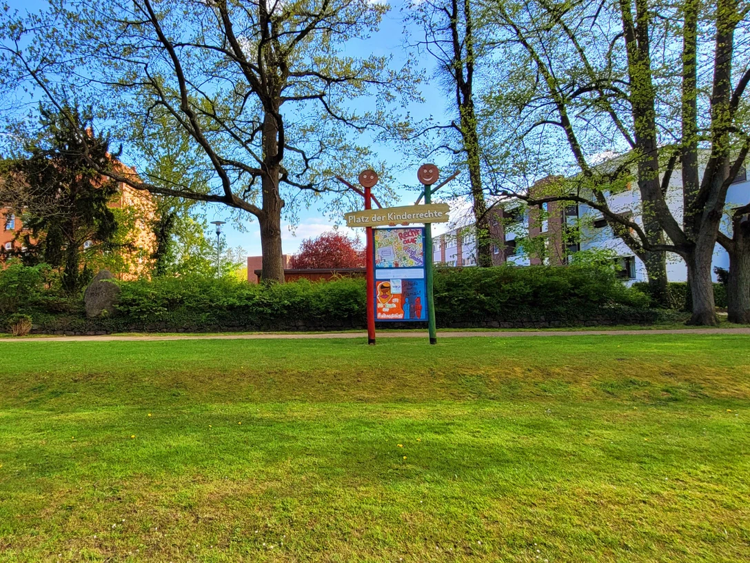Platz der Kinderrechte_1980x1080_Silvana Dust_20230510 (3).jpg Auf der Wiese steht ein Schild mit ‚Platz der Kinderrechte‘. Dahinter sind Bäume und Häuser zu sehen, viel Platz zum Spielen