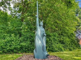 Eine kantige Statue von Hermann dem Cherusker erhebt sich vor dichtem grünem Laubwerk.