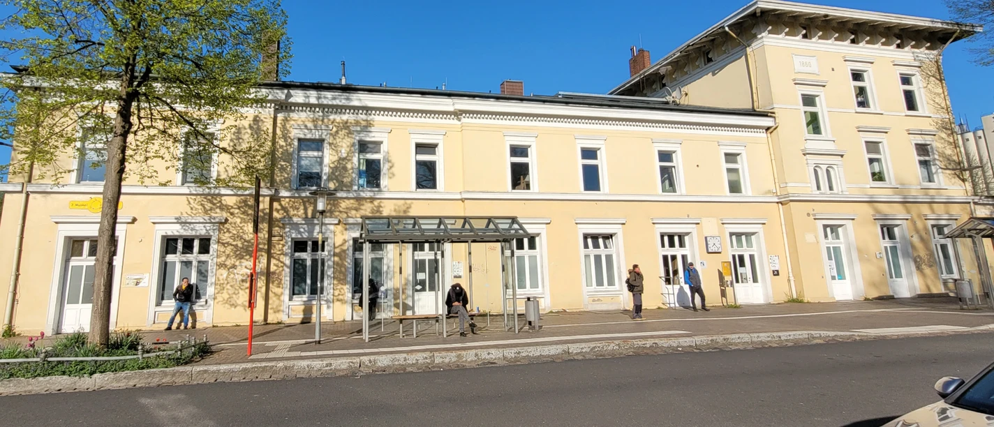 Bahnhof Ratzeburg_1980x1080_Silvana Dust_20230510 (2).jpg Leicht gelb gestrichenes Bahnhofsgebäude mit weißen Fenstern. Vor dem Gebäude ist eine Bushaltestelle