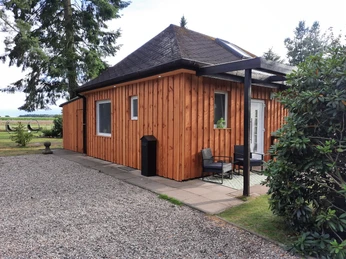 Holzverkleidetes Ferienhaus mit steilem Dach, umgeben von Bäumen und Kiesweg, lädt zur Erholung ein.