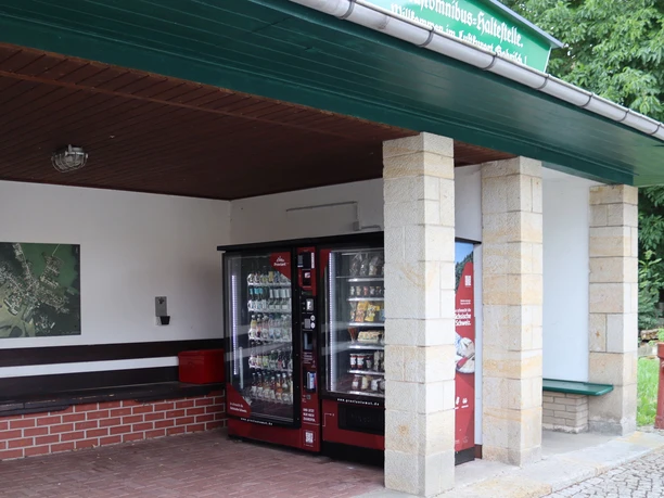 Overdekte bushalte met rode drank- en snackautomaat, een bankje en een wandkaart ernaast; rustige, landelijke sfeer.