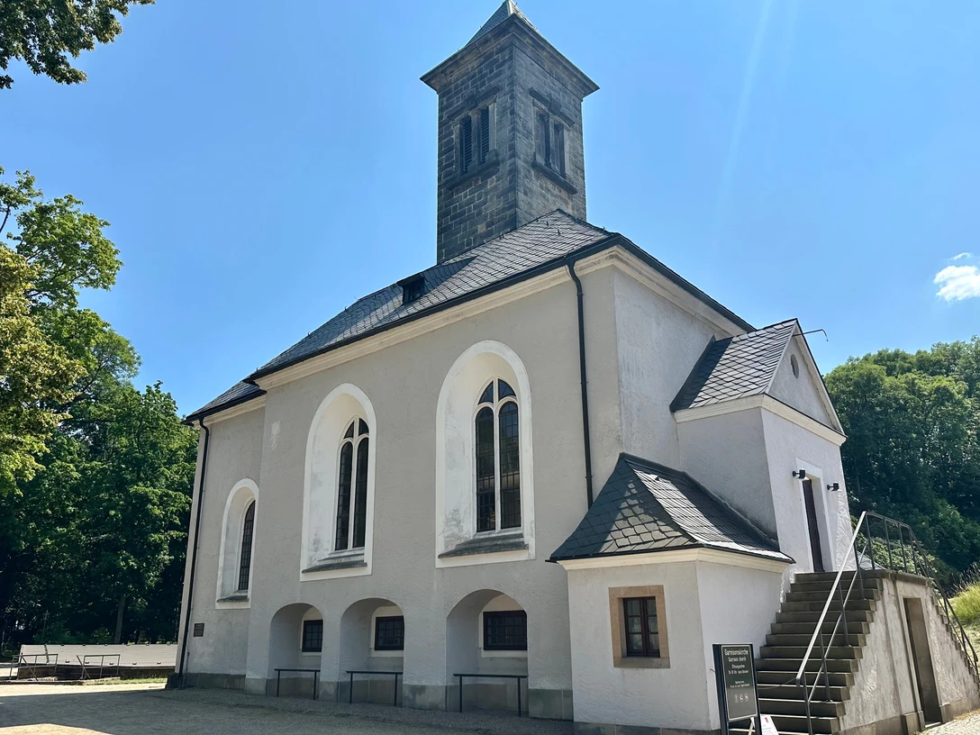 Garnisionskirche auf der Festung Königstein Weiße Kirche mit grauem Turm und Spitzdach, umgeben von Bäumen unter klarem, blauem Himmel; ruhige, sonnige Atmosphäre.