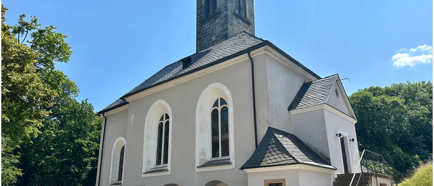 Garnisionskirche auf der Festung Königstein White church with gray tower and pointed roof, surrounded by trees under a clear blue sky; quiet, sunny atmosphere.