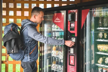 Proviantomat in Rathen Ein Mann mit Rucksack bedient einen Getränke- und Snackautomaten, der mit Flaschen und verpackten Lebensmitteln gefüllt ist.