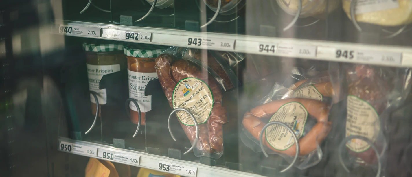 Vending machine with various food products, including sausages, soups in a jar and snacks, on a shelf with price tags.