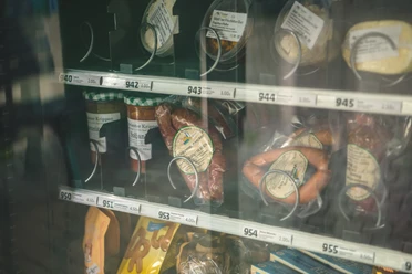 Proviantomat Verkaufsautomat mit verschiedenen Lebensmitteln, darunter Wurstwaren, Suppen im Glas und Snacks, in einem Regal mit Preisschildern.