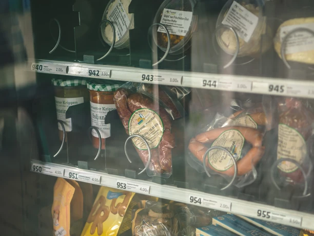 Proviantomat Verkaufsautomat mit verschiedenen Lebensmitteln, darunter Wurstwaren, Suppen im Glas und Snacks, in einem Regal mit Preisschildern.