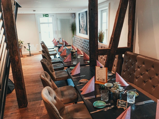 ManuFaktur Speisekarte Herbst 2021-31.jpg Gemütliches Restaurant mit langer, gedeckter Tafel, braunen Ledersesseln und Holzbalken an den Wänden.