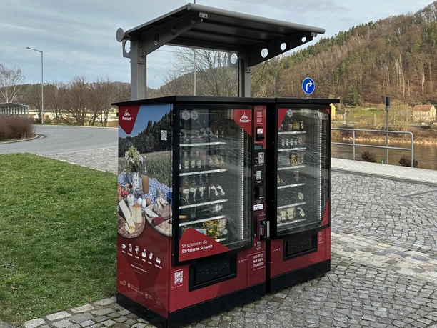Červený automat s regionálními produkty, obklopený dlážděným náměstím a zelenou loukou, s řekou a zalesněnými kopci v pozadí.