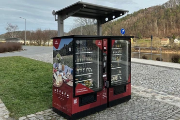 Proviantomat in Bad Schandau Roter Verkaufsautomat mit regionalen Produkten, umgeben von gepflastertem Platz und grüner Wiese, im Hintergrund Fluss und bewaldete Hügel.