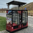 Proviantomat in Bad Schandau Roter Verkaufsautomat mit regionalen Produkten, umgeben von gepflastertem Platz und grüner Wiese, im Hintergrund Fluss und bewaldete Hügel.Red vending machine with regional products, surrounded by a paved square and green meadow, with a river and wooded hills in the background.Červený automat s regionálními produkty, obklopený dlážděným náměstím a zelenou loukou, s řekou a zalesněnými kopci v pozadí.Czerwony automat z produktami regionalnymi, otoczony wybrukowanym placem i zieloną łąką, z rzeką i zalesionymi wzgórzami w tle.Rode automaat met regionale producten, omgeven door een geplaveid plein en groene weide, met een rivier en beboste heuvels op de achtergrond.Distributore automatico rosso di prodotti regionali, circondato da una piazza lastricata e da un prato verde, con un fiume e colline boscose sullo sfondo.