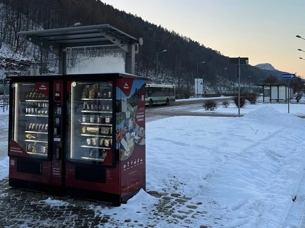 Červený automat stojí na sněhu na autobusové zastávce, obklopen zimní krajinou a projíždějícím autobusem.