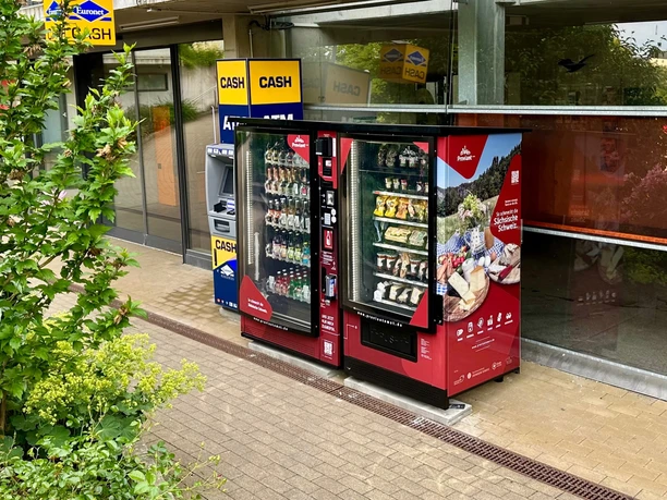 Twee rode automaten met snacks en drankjes staan naast een geldautomaat voor een gebouw omringd door groene vegetatie.