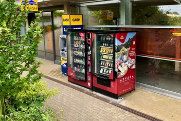 Proviantomat am Parkhaus der Festung Königstein Zwei rote Verkaufsautomaten mit Snacks und Getränken stehen neben einem Geldautomaten vor einem Gebäude, umgeben von grüner Vegetation.