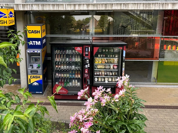 Geldautomaat en twee snackautomaten voor een gebouw met een glazen gevel, omringd door groene planten en roze bloemen.