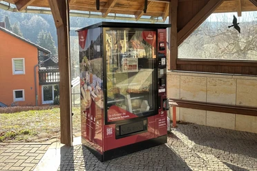 Proviantomat in Leupoldishain Ein roter Verkaufsautomat mit der Aufschrift "So schmeckt die Sächsische Schweiz" steht in einem überdachten Bereich, im Hintergrund ein orangefarbenes Haus.