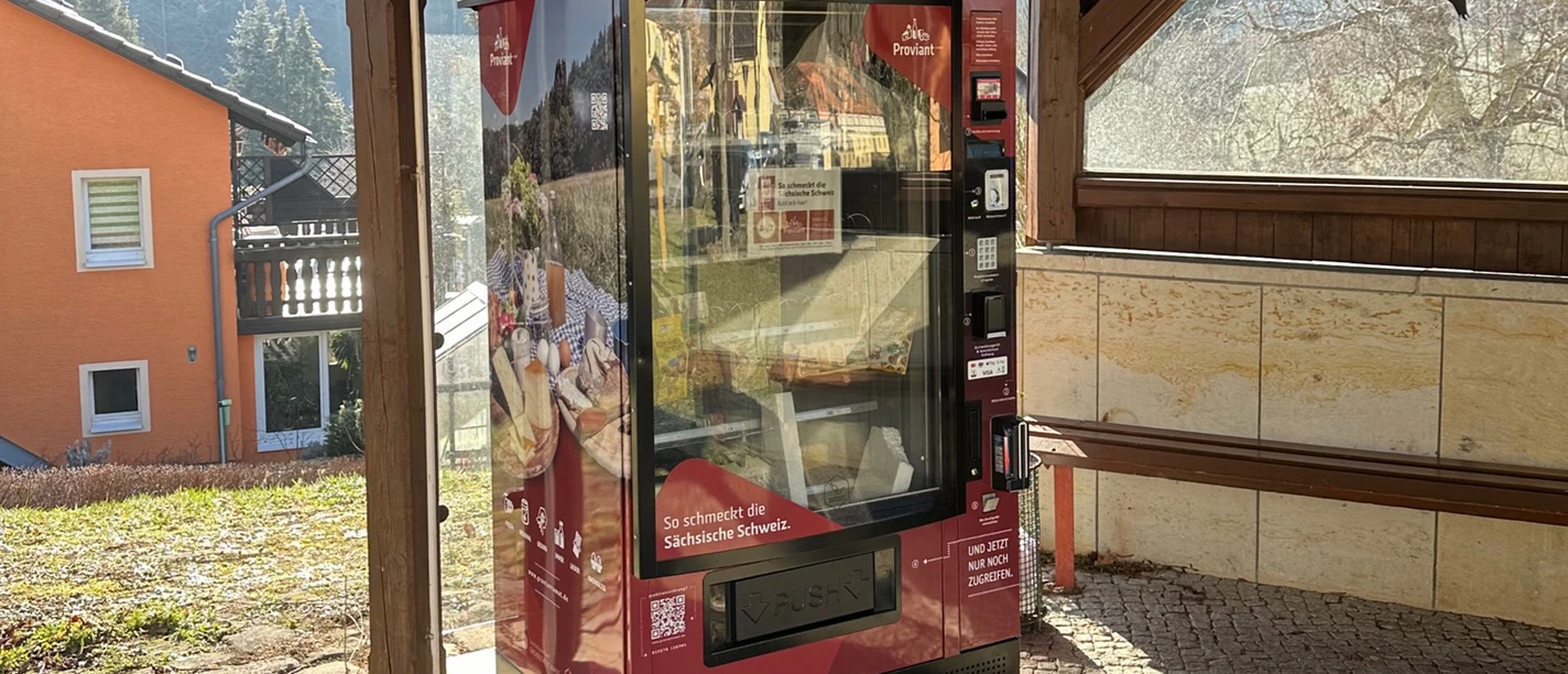 Proviantomat in Leupoldishain Ein roter Verkaufsautomat mit der Aufschrift "So schmeckt die Sächsische Schweiz" steht in einem überdachten Bereich, im Hintergrund ein orangefarbenes Haus.