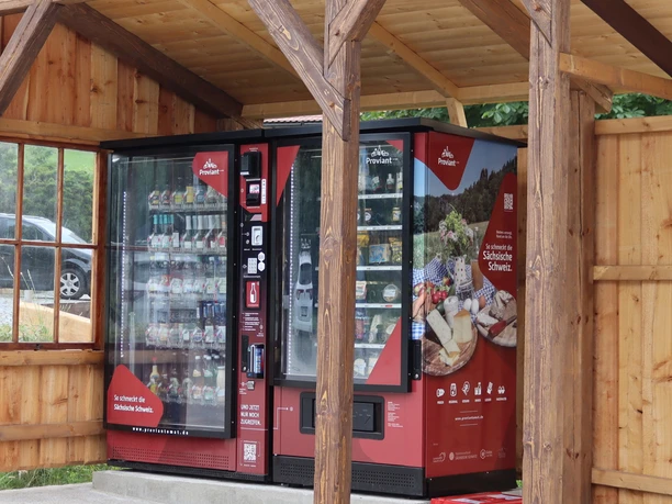Rode automaat met hapjes en drankjes onder een houten afdak, omgeven door een landelijke omgeving.