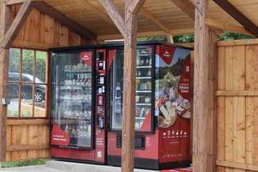 Proviantomat in Pfaffendorf Roter Verkaufsautomat mit Snacks und Getränken unter einem hölzernen Unterstand, umgeben von ländlicher Umgebung.