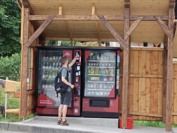 Een man met een rugzak bedient een rode drank- en snackautomaat onder een houten afdak.