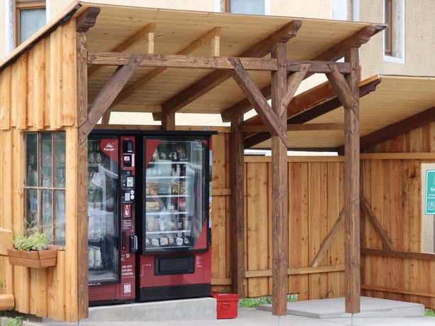 Een rode automaat staat onder een houten afdak naast een gebouw, omringd door houten muren en een bord met de tekst "Playground".