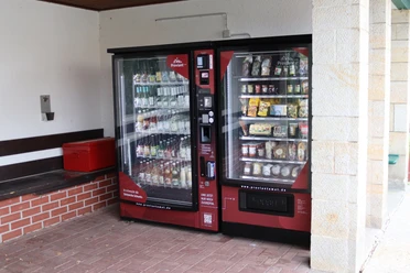 Two red vending machines stand next to each other; the one on the left offers drinks, the one on the right snacks and food. Next to it is a red cool box.