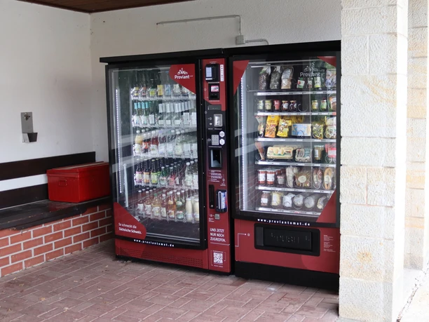 Twee rode automaten staan naast elkaar; de linker biedt drankjes, de rechter snacks en eten. Ernaast staat een rode koelbox.