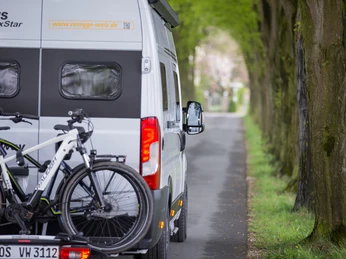 Ein Wohnmobil mit Fahrradträger fährt auf einer von Bäumen gesäumten Landstraße entlang.