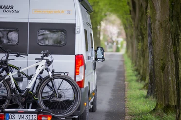 Camperplaatsen in de regio Osnabrück Een camper met fietsendrager rijdt over een door bomen omzoomde landweg.