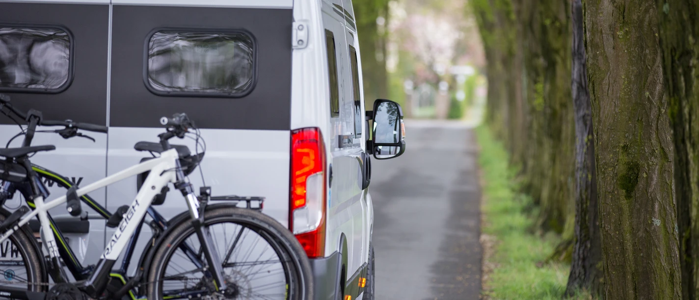 Wohnmobilstellplätze im Osnabrücker Land Ein Wohnmobil mit Fahrradträger fährt auf einer von Bäumen gesäumten Landstraße entlang.