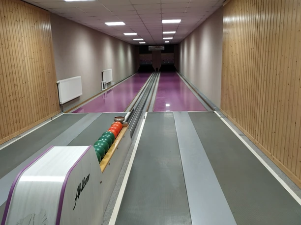 An empty bowling alley with gray and pink lanes, green and orange balls on the left; wooden walls and ceiling lighting.