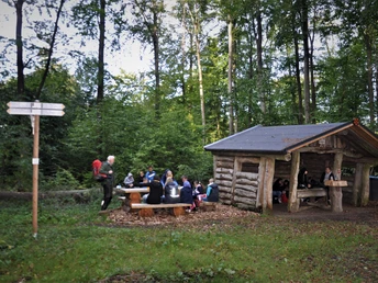 Wanderer in einem waldigen Rastplatz mit Blockhütte und Picknicktischen im Grünen.