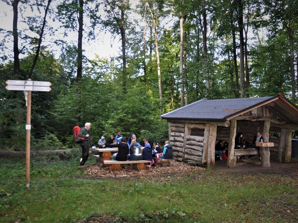Blockhütte_BlombergMarketing Wanderer in einem waldigen Rastplatz mit Blockhütte und Picknicktischen im Grünen.