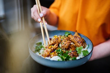Eine Schale mit asiatischem Essen wird mit Stäbchen gehalten. Der Inhalt besteht aus Fleisch, Gemüse und Reis.