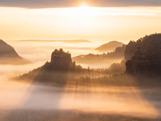 Blick auf den Winterstein (Hinteres Raubschloss) Sonnenaufgang über nebelverhangenen Bergen mit Felsen und Bäumen, die von warmem, goldenem Licht durchflutet werden.