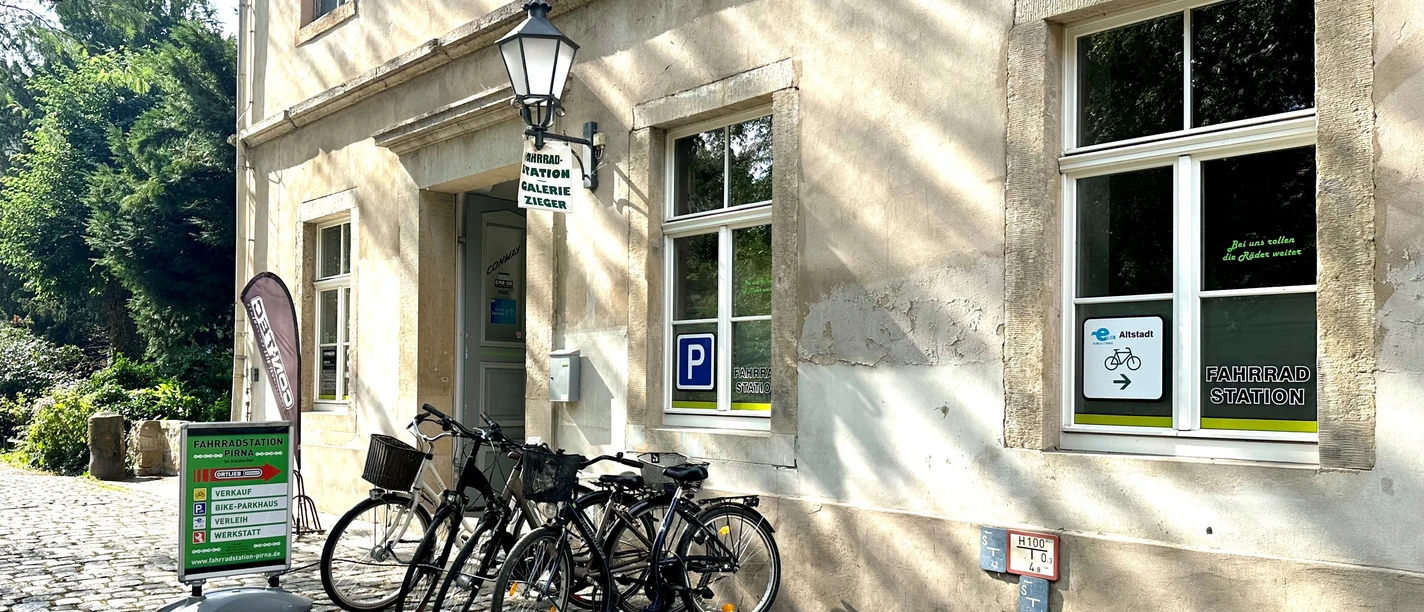 Fahrradstation Pirna Fahrradstation vor einem historischen Gebäude mit mehreren geparkten Fahrrädern, Schildern und einem grünen Hinweisschild auf Kopfsteinpflaster.