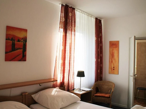 Ein gemütliches Schlafzimmer mit einem Doppelbett und warmen Farben. An den Wänden hängen zwei Bilder, und das Fenster ist mit roten Vorhängen geschmückt.