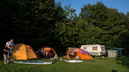 Camping 3 ©Jens Butz.jpg Auf dem Campingplatz stehen mehrere Zelte und ein Wohnanhänger, umgeben von Personen, die sich im Freien aufhalten