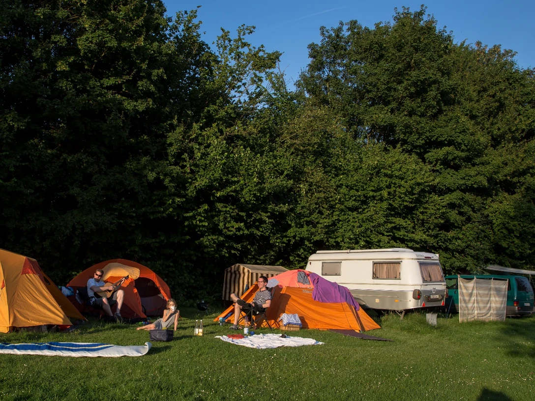 Camping 3 ©Jens Butz.jpg Auf dem Campingplatz stehen mehrere Zelte und ein Wohnanhänger, umgeben von Personen, die sich im Freien aufhalten