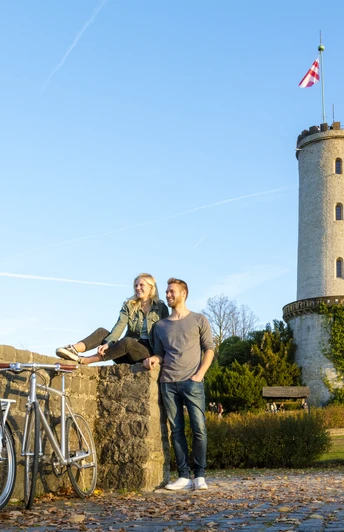 Zwei Personen sitzen auf einer Mauer vor einem mittelalterlichen Turm und genießen die Herbstlandschaft.