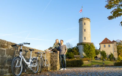 Bielefelder Romanzen_Sparrenburg_Ulrich Helweg.jpg Zwei Personen sitzen auf einer Mauer vor einem mittelalterlichen Turm und genießen die Herbstlandschaft.