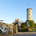 Bielefelder Romanzen_Sparrenburg_Ulrich Helweg.jpg Zwei Personen sitzen auf einer Mauer vor einem mittelalterlichen Turm und genießen die Herbstlandschaft.